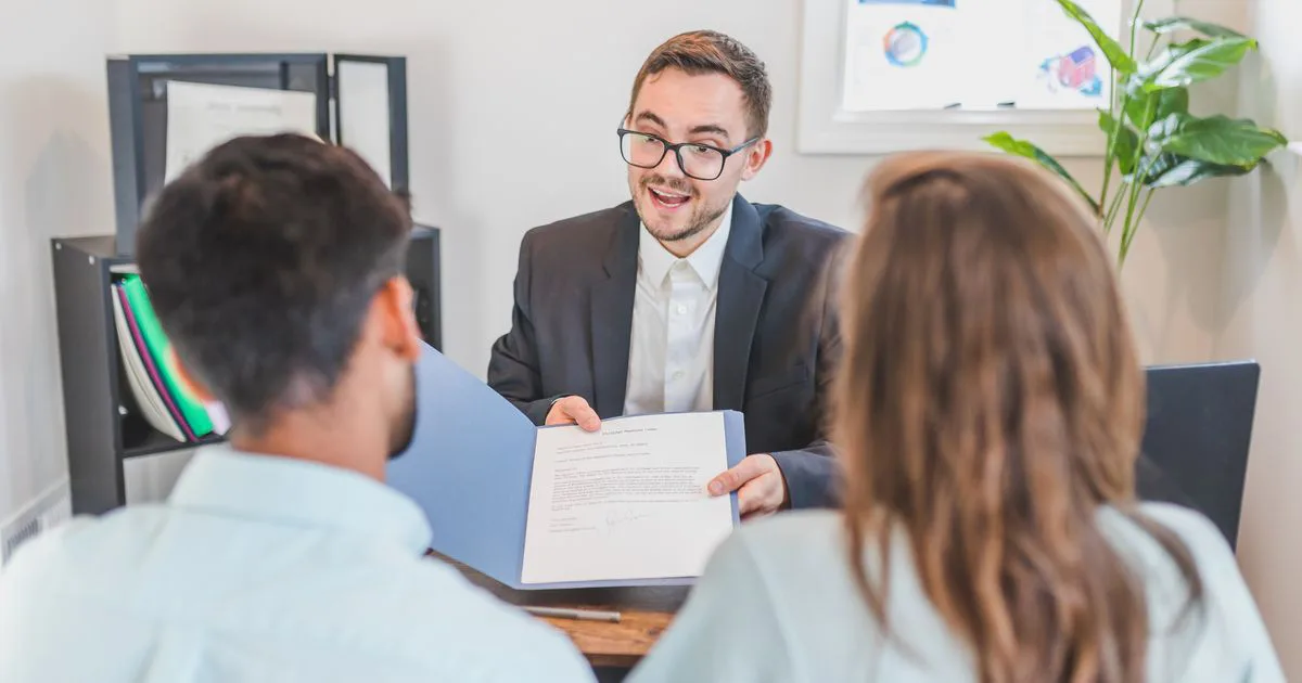 A broker holds a contract open across the desk while a couple sits opposite, hearing the terms explained before anything is signed.