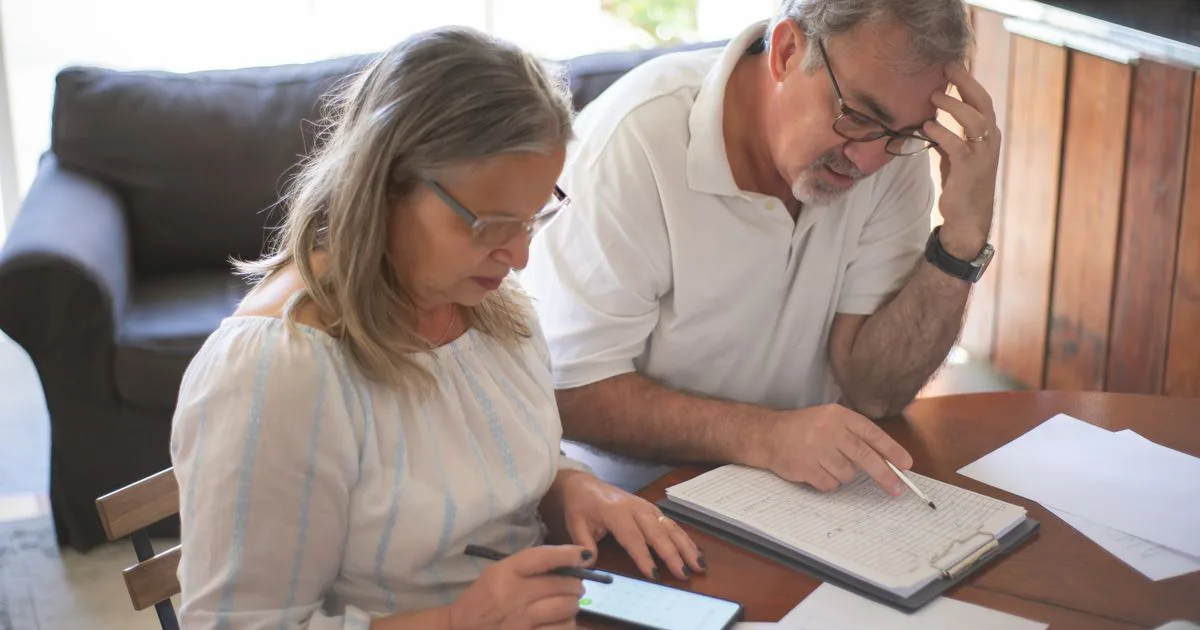 A retirement-age couple sits at their table at home, working through paperwork together with a pen and a phone calculator.