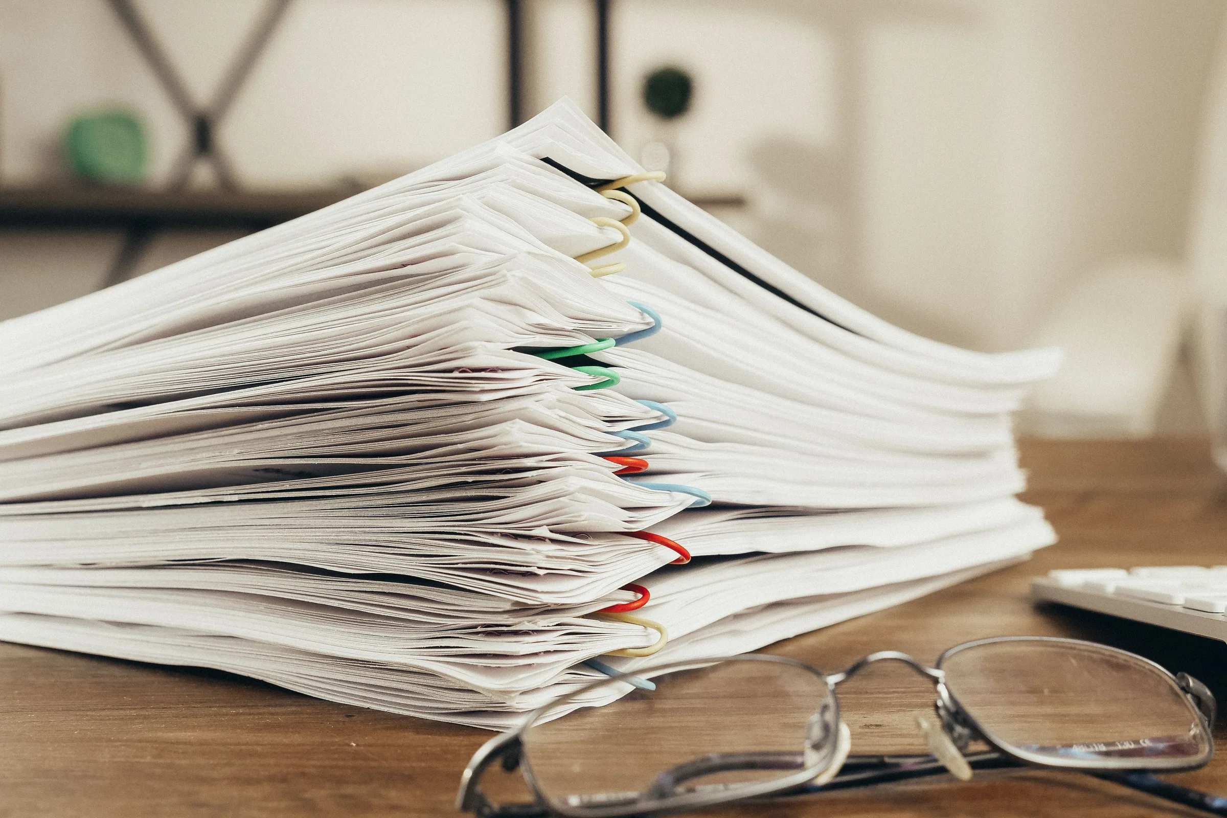 A thick stack of paperwork gathered in coloured plastic clips on a timber desk, with reading glasses resting in front of it