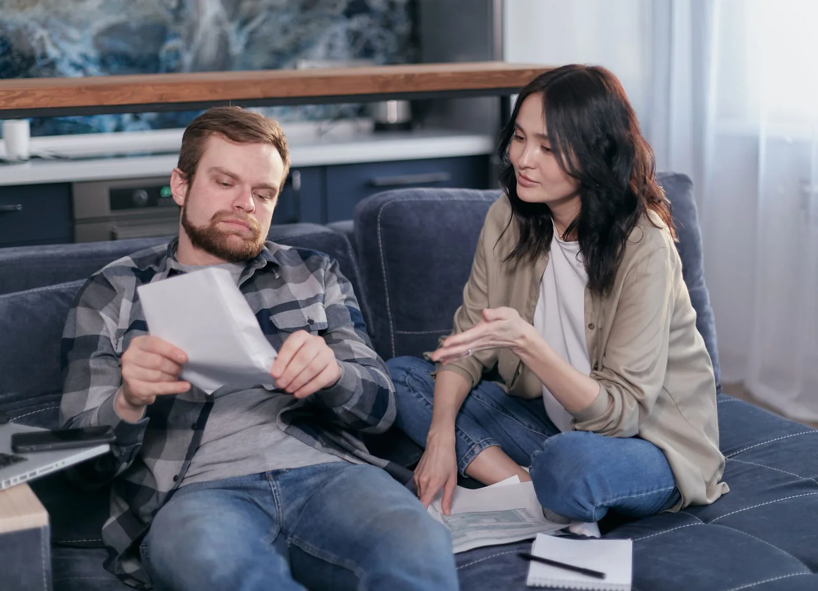 A young couple sits together on their living-room sofa working through property paperwork, one holding a statement while the other points to a figure, with a laptop beside them.