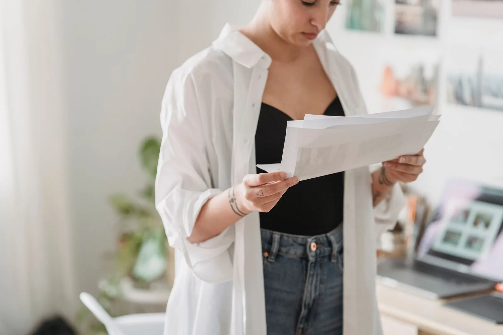 A young woman stands in her home reading through printed documents, checking the details against the FHSS eligibility rules with a laptop open on the desk behind her.