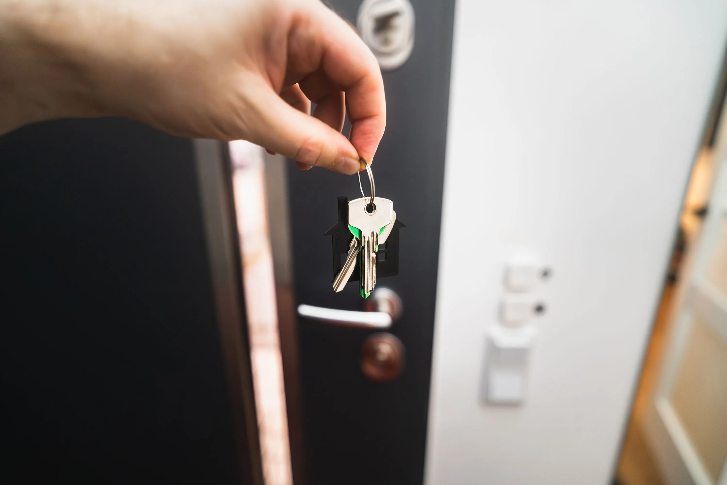 A hand holds a set of keys with a small house-shaped keychain in front of an open front door, just after settlement on a first home.