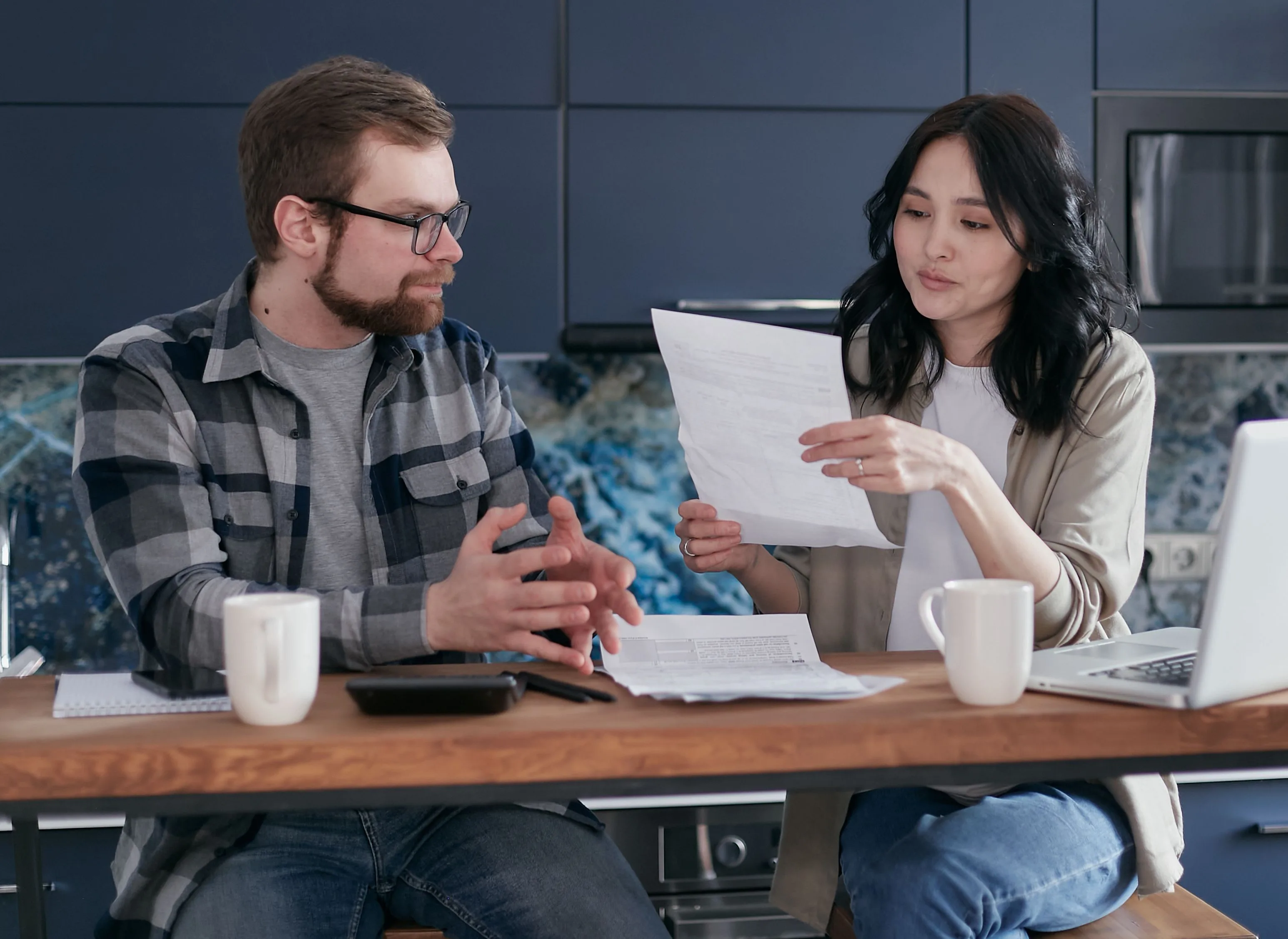 A young couple sits at their kitchen table reviewing super statements and paperwork together, with a laptop, a calculator and two coffee mugs in front of them.