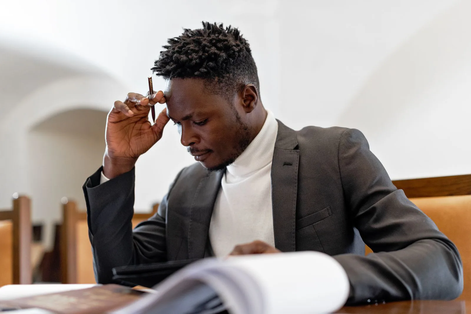 A young man pauses with a pen pressed to his forehead as he reads through a stack of paperwork, thinking carefully before making a financial decision.
