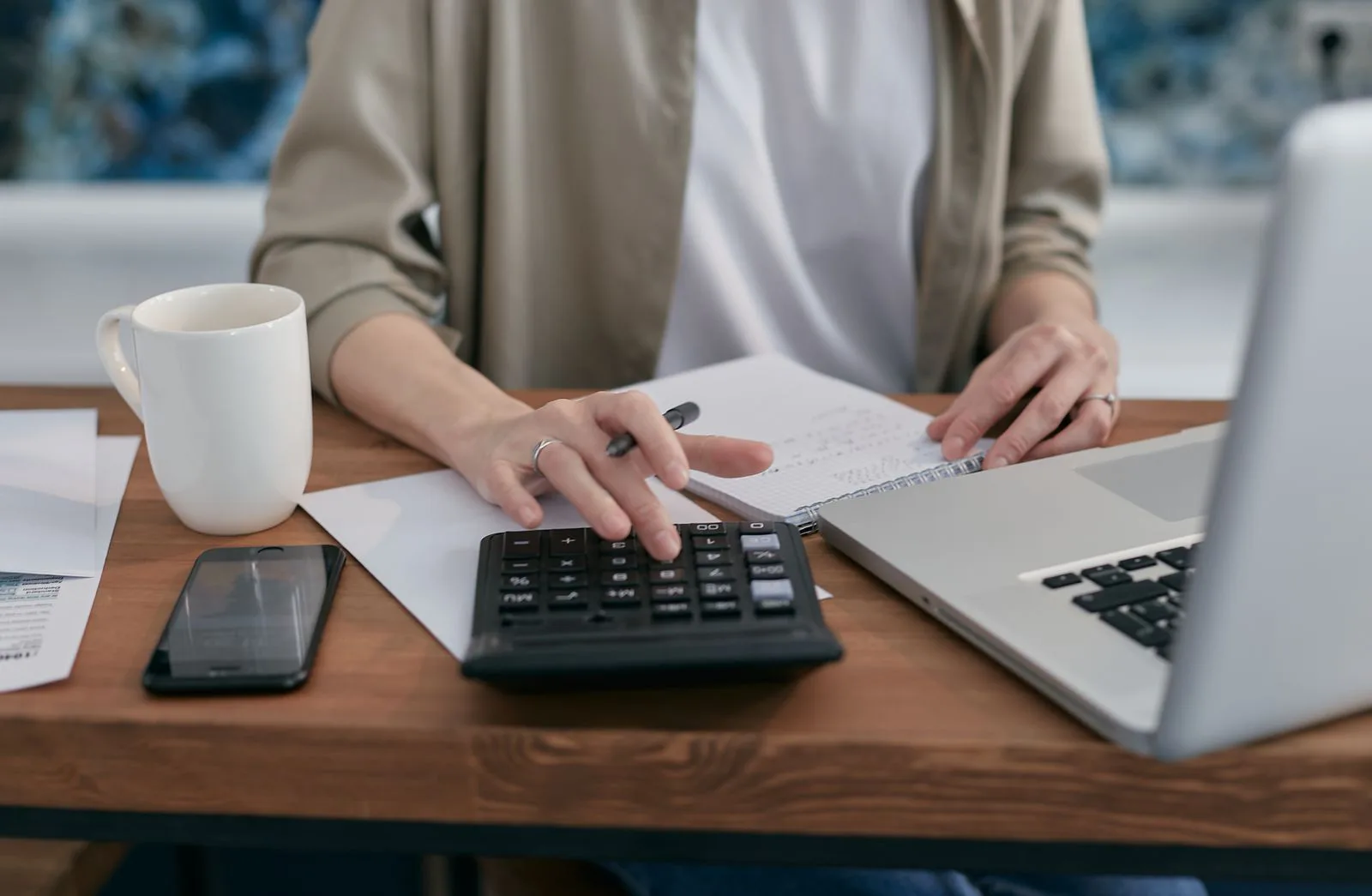A young person sits at a wooden desk working out figures on a calculator, with a notepad, an open laptop and a coffee mug beside them, weighing up whether the numbers stack up.